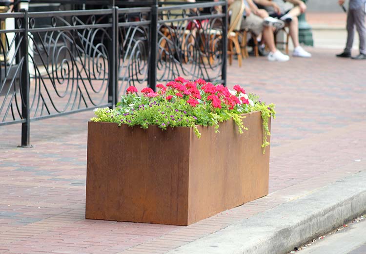 Low, rectangular Form and Fiber planter box on a brick sidewalk near the curb. An outdoor cafe is visible just beyond the sidewalk.