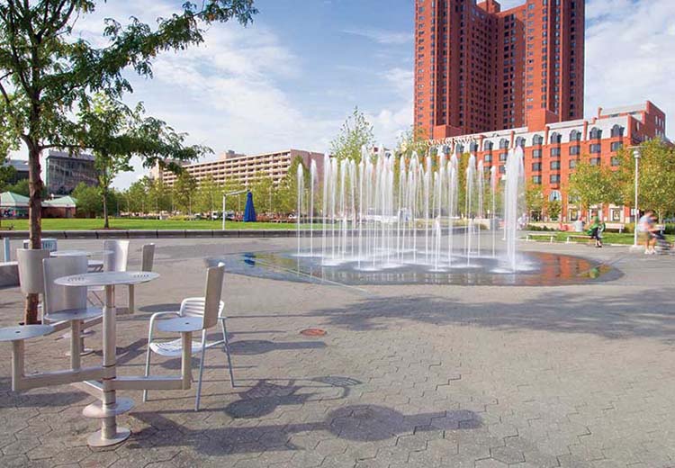 A view of West Shore Park, looking out across the fountain. Tall, red brick buildings are in the background. A table and chairs are in the foreground. The ground is made of Hanover asphalt hexagonal block pavers.