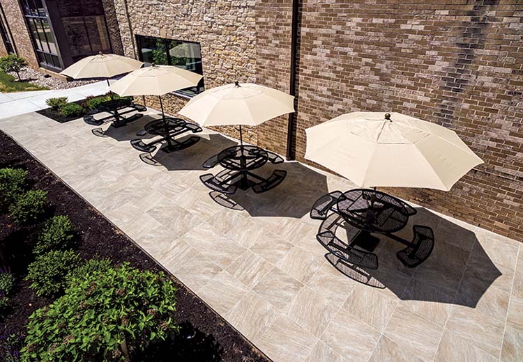 Outdoor seating area for employees. Near the building wall are 4 tables with chairs with umbrellas at each table. The ground is Hanover porcelain pavers in a York pattern, meant to look like natural stone