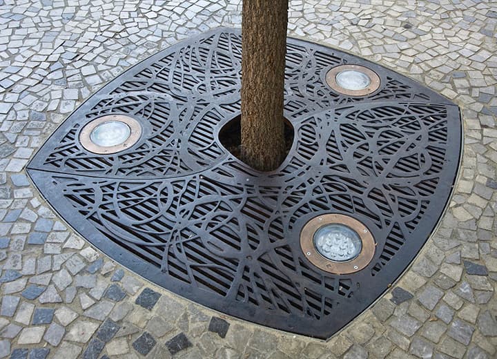A tree trunk with a custom decorative metal grate by Ironsmith around the roots. Three uplights are embedded into the grate.