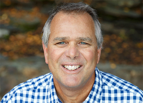 Head and shoulders portrait of a man smiling at the camera. He is wearing a checkered shirt and is greying at the temples.