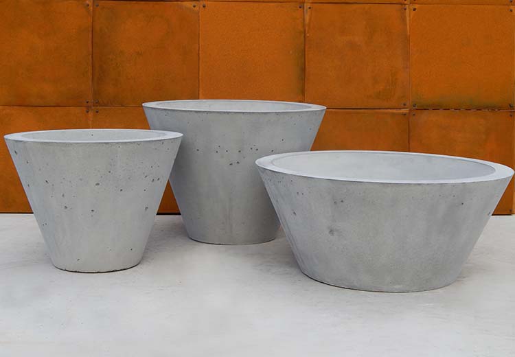 Three sizes of Form and Fiber Cypress round flared planters in concrete planters in front of a terracotta stone wall