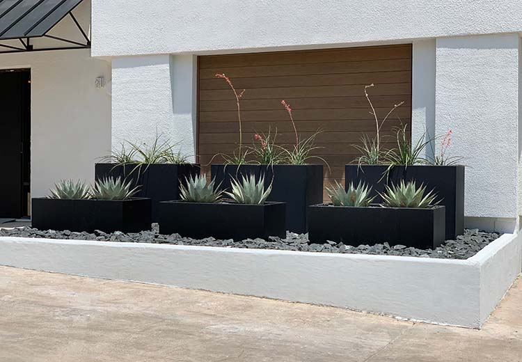 Two rows of Form and Fiber custom steel planters in a rock garden in front of a stucco office building