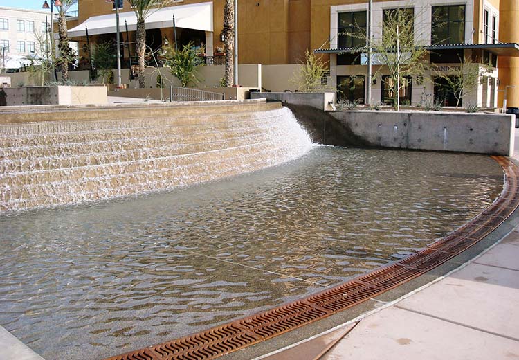 A fountain at an outdoor shopping mall with Ironsmith Conquistador-style trench grates at the sidewalk edge to funnel away excess water.