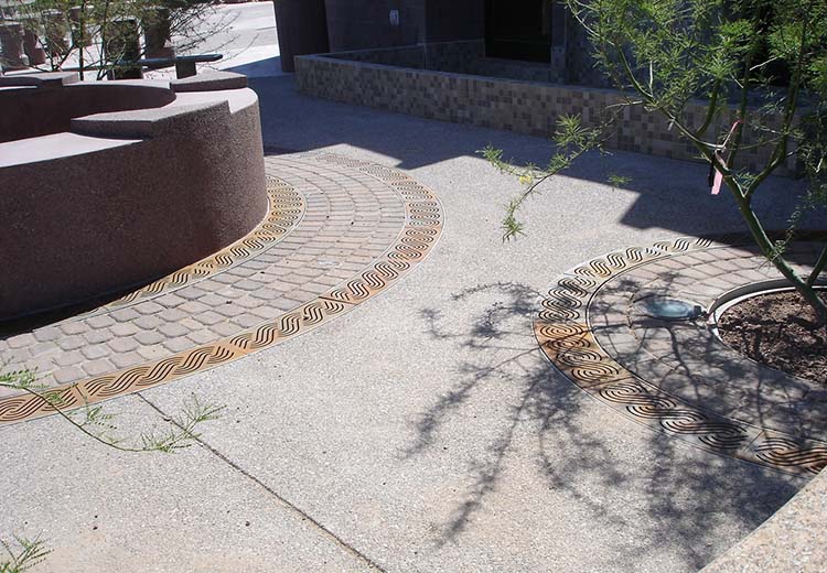 Outdoor area with a short curved wall. Around the base of the curved wall there are two rings of Ironsmith Marina-style trench grating with 3 rows of bricks between them.