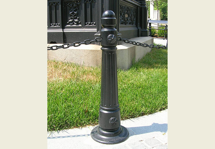 Ironsmith Victorian-style black metal bollard in front of a monument. A chain links the bollard to others to form a barrier