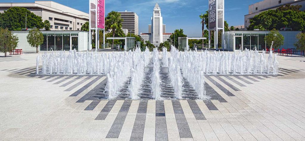 Multi-spout fountain placed in a paved open courtyard. The Hanover Prest pavers make stripes. The Los Angeles Music Center is in the distance.